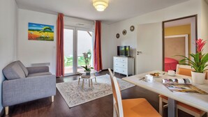 Una sala de estar moderna con un sofá, una mesa de comedor con sillas, un televisor y un ventanal corredero que da a un espacio exterior.