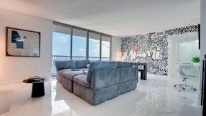A modern living room with a grey sofa, a black coffee table, and a large window offering a view of the sky. - The Noir Suite 3BR 2BTH Glam Room PS5 Pool Gym (Miami)