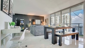 A modern living room with a grey sofa, a wooden dining table, and a large window offering a view of high-rise buildings. - The Noir Suite 3BR 2BTH Glam Room PS5 Pool Gym (Miami)
