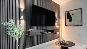 A modern living room with a large flat-screen TV mounted on a wooden panel wall, a small side table with a decorative item, and a potted plant. - The Noir Suite 3BR 2BTH Glam Room PS5 Pool Gym (Miami)