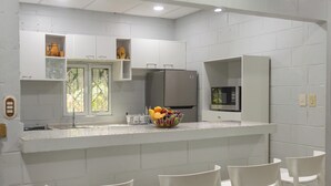 A modern kitchen with white cabinets, a stainless steel refrigerator, a microwave, and a fruit bowl on the counter. There are four white bar stools around the counter.