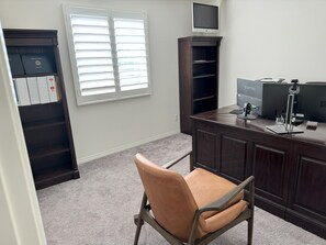 Ein Homeoffice mit Schreibtisch, Computer und Stuhl. Ein Fenster mit Jalousien, ein Bücherregal und ein Teppichboden sind ebenfalls vorhanden.