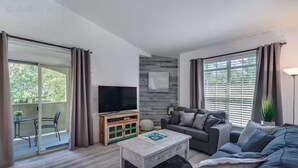 Ein modernes Wohnzimmer mit Sofa, Couchtisch und Fernseher. Es gibt einen Balkon mit Tisch und Stühlen sowie eine Holzkommode mit einem Fernseher.