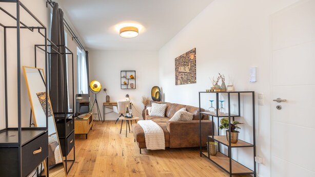 A modern, well-lit room with a wooden floor, a brown leather sofa, a desk with a lamp, and a bookshelf. - Studio Augusta: modern, fully equipped, Rhine 50m, business & private, all-inclusive (Koblenz)