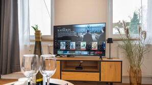A hotel room with a TV displaying a series titled "Die Kaiserin," a wooden TV stand, a vase with dried plants, and a tray with wine glasses and candles. - Studio Augusta: modern, fully equipped, Rhine 50m, business & private, all-inclusive (Koblenz)