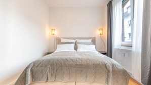 A neatly made bed with a quilted cover, two bedside lamps, and a window with curtains. - Studio Augusta: modern, fully equipped, Rhine 50m, business & private, all-inclusive (Koblenz)