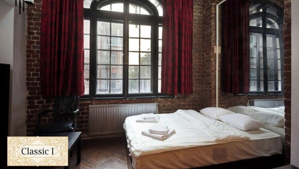 Habitación clásica con pared de ladrillo, dos amplias ventanas en arco con cortinas rojas, una cama con ropa de cama blanca y un radiador debajo de las ventanas.