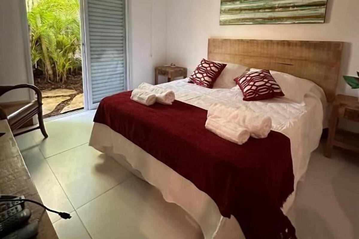 A bedroom with a bed, a nightstand, a window with a view of greenery, and a door leading outside.