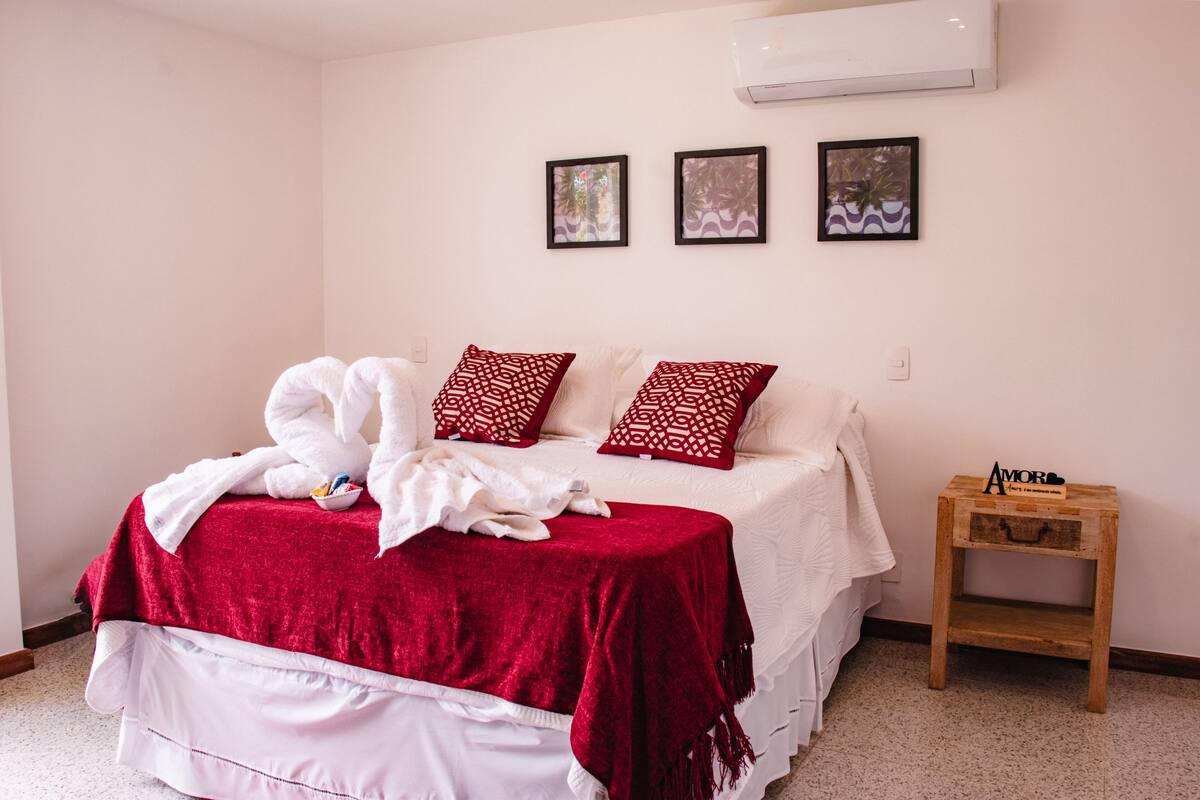 A bedroom with a bed, red and white pillows, a red blanket, a white robe, and a small wooden nightstand with a decorative item.