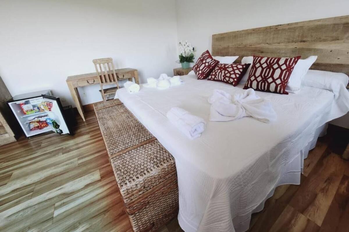 A hotel room with a large bed, a wooden headboard, a small table, a wicker bench, and a minibar.