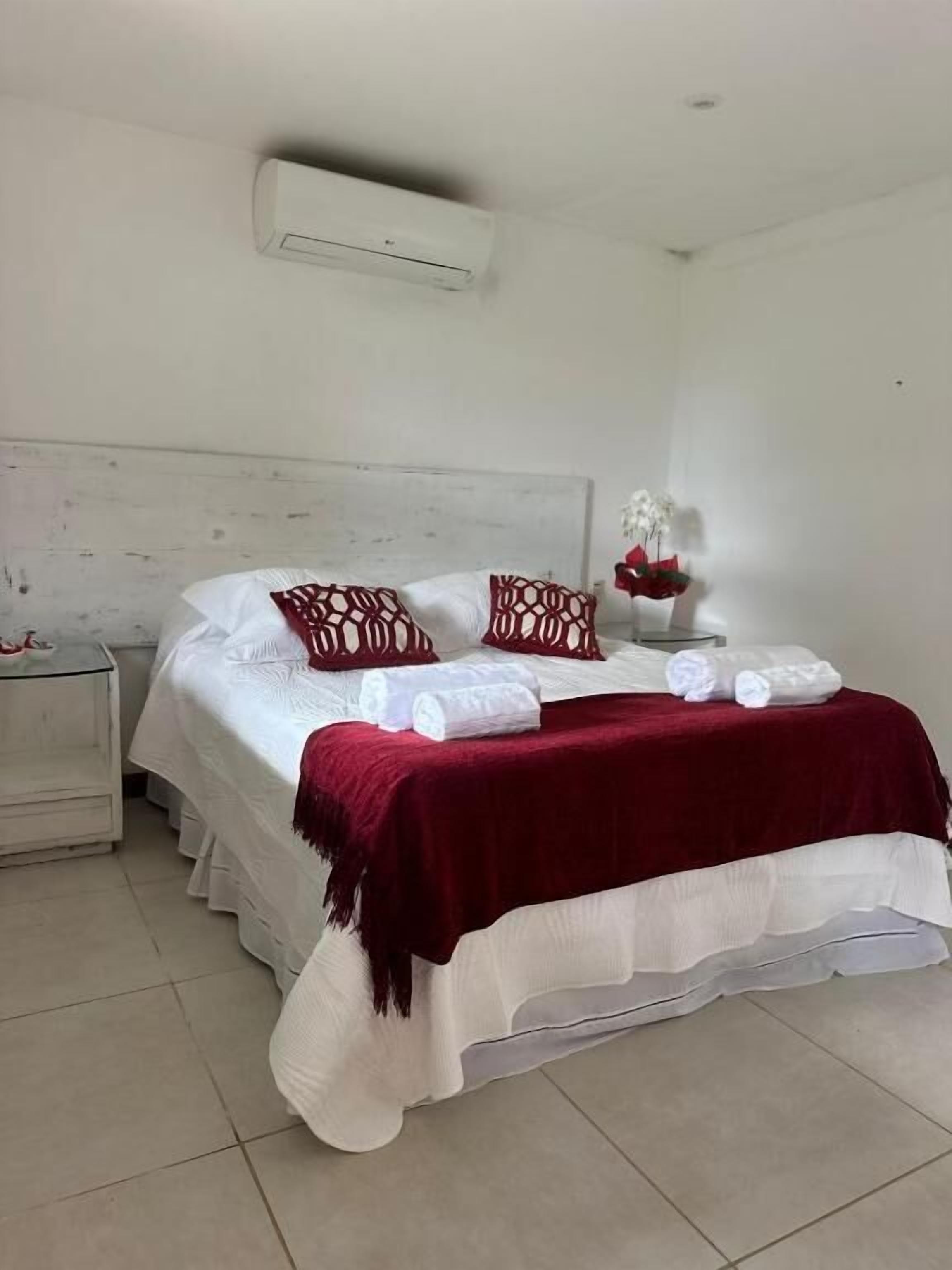 A neatly made bed with white linens and red decorative pillows, a red throw blanket, and a white vase with flowers on the nightstand beside it.