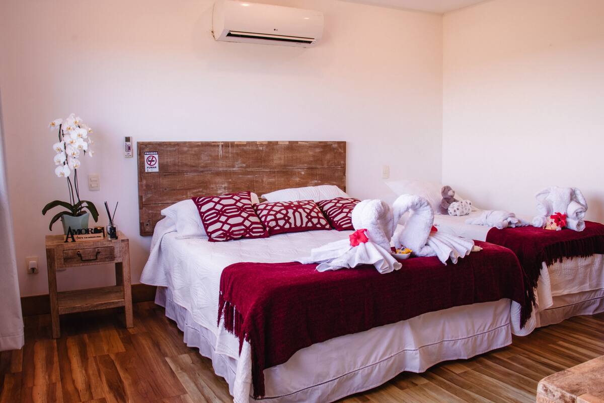 A hotel room with two beds, a wooden headboard, a red throw blanket, and a small bedside table with a plant and a sign.