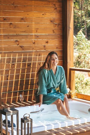 A person sitting in a wooden cabin with a hot tub, surrounded by wooden beams and greenery.