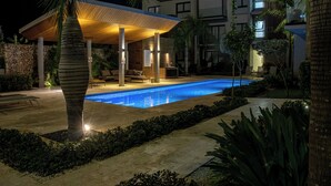 A nighttime swimming pool area with a covered seating area, palm trees, and a stone wall. - Cap Cana Luxury 2 bedroom and 2 bath condo, Pool, near Juanillo beach, Golf




 (Las Canas)