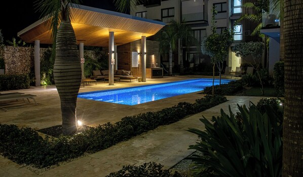 A nighttime swimming pool area with a covered seating area, palm trees, and a stone wall.