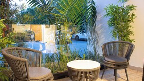 A patio with wicker chairs and a small round table, surrounded by lush greenery and a view of a car and building in the background. - Cap Cana Luxury 2 bedroom and 2 bath condo, Pool, near Juanillo beach, Golf




 (Las Canas)