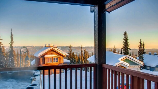 A wooden cabin with a hot tub on a balcony overlooking a snowy landscape with trees and distant mountains. - 385 Moonshine Crescent by Okanagan Premier (Beaverdell)
