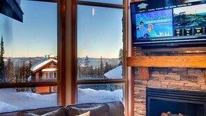 A cozy living room with a large TV displaying a skiing program, a stone fireplace, and a view of a snowy mountain landscape. - 385 Moonshine Crescent by Okanagan Premier (Beaverdell)
