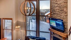 A modern interior with a large window, a stone wall, and a flat-screen TV displaying a weather forecast. - 385 Moonshine Crescent by Okanagan Premier (Beaverdell)