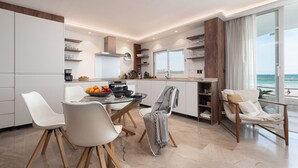 Cozinha moderna com armários brancos, área de refeições com mesa de vidro e vista para a praia através de grandes janelas.