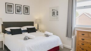 Ein ordentlich bezogenes Bett mit Kissen und gerollten Handtüchern, ein Nachttisch mit Lampe, eine Kommode mit Schubladen und gerahmte Bilder an der Wand.