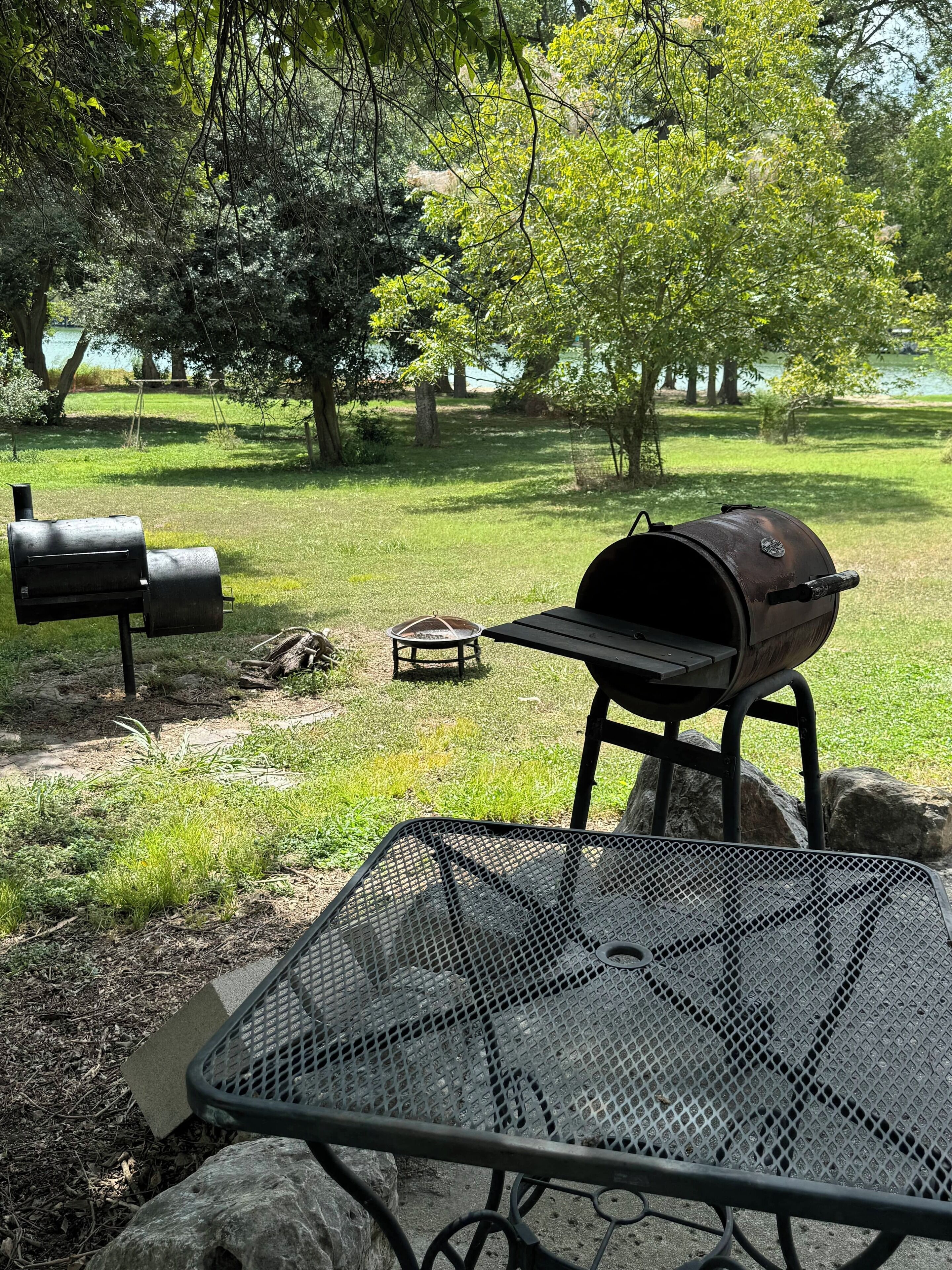 Una parrilla y una mesa de metal en un espacio al aire libre con césped, árboles y un lago al fondo.