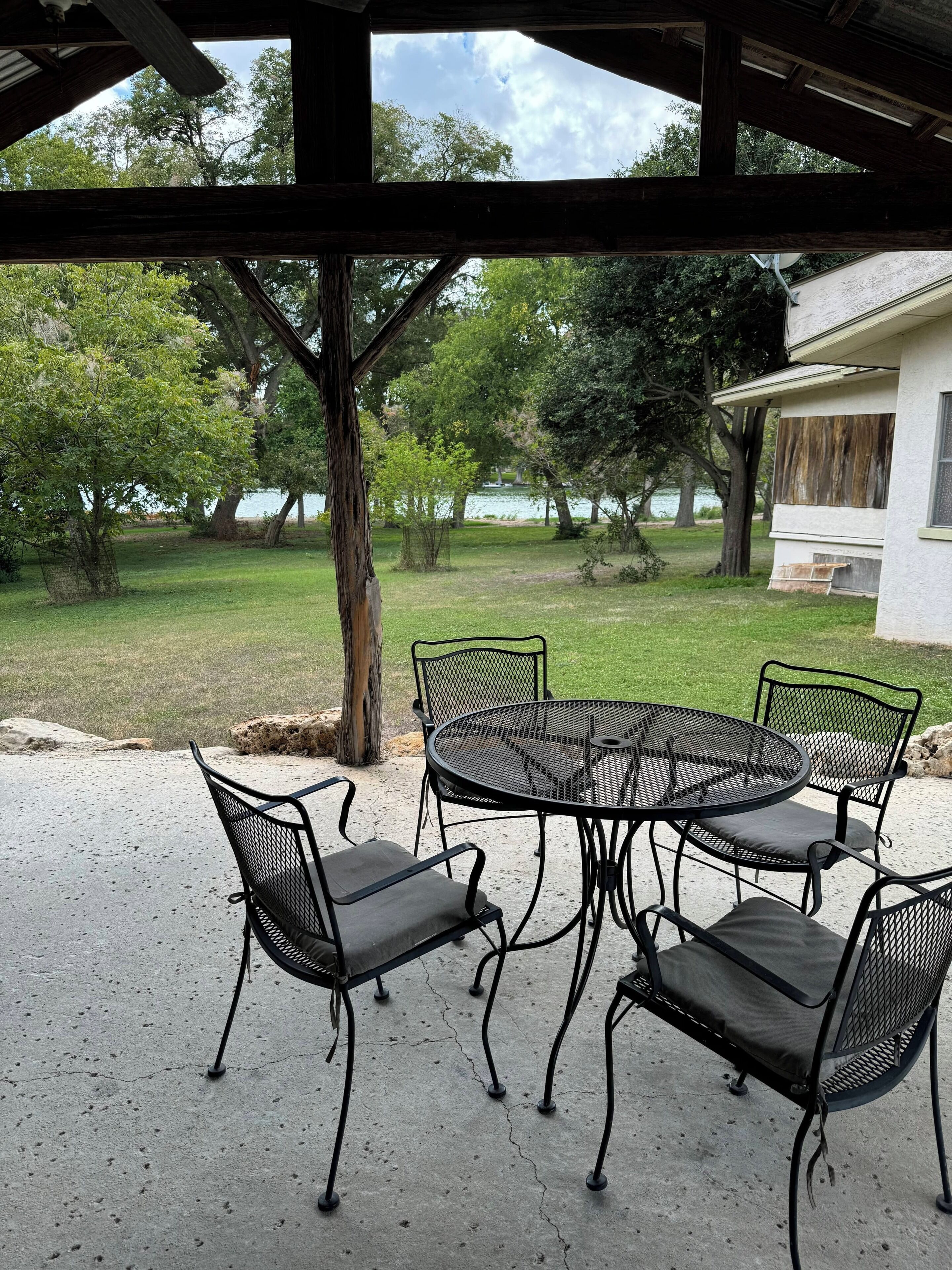 A covered patio with a round metal table and four chairs, overlooking a grassy area with trees and a lake.
