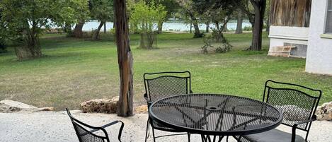 A covered patio with a round metal table and four chairs, overlooking a grassy area with trees and a lake.