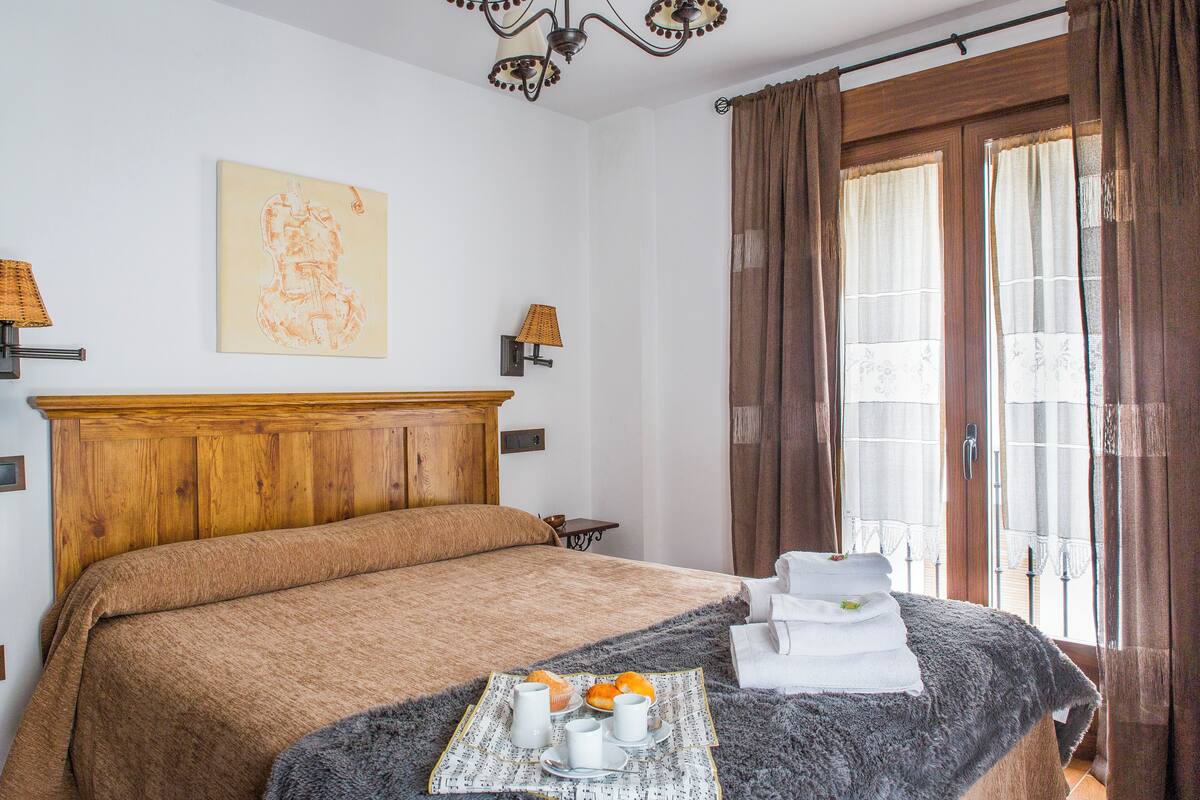 Un lit bien fait, avec une tête de lit marron, une table de chevet sur laquelle se trouve un plateau avec des articles pour le petit-déjeuner, et une fenêtre avec des rideaux.