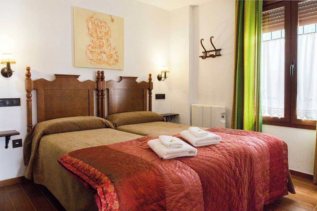 Un lit double avec une couette rouge et marron, deux tables de chevet avec des lampes, un tableau au mur et une fenêtre avec des rideaux verts.