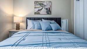Una cama bien tendida con edredón azul y blanco, una lámpara de noche y un cuadro de una escena callejera en la pared.
