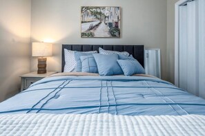 Un letto rifatto con cura, coperto da un piumone blu e bianco, una lampada da comodino e un quadro raffigurante una scena di strada al muro.