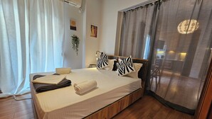 A neatly made bed with towels, a headboard with decorative pillows, a wooden floor, and a view of a dining area through a window.