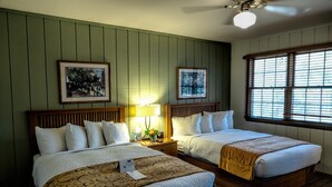 Quarto de hotel com duas camas, cabeceira de madeira, mesa de cabeceira com abajur, quadros nas paredes e janela com persianas.