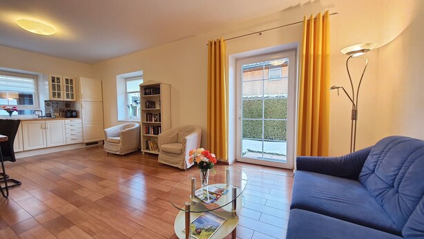 A living room with a sofa, a coffee table with magazines, a bookshelf, a chair, and a kitchen area with cabinets and a window. - Comfortable, very quiet and sunny apartment. Only 5 minutes to the Silbersee (Villach)