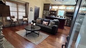 Ein geräumiges Wohnzimmer mit einer dunkelbraunen Ledersofa, einem Couchtisch und zwei Sesseln. Im Hintergrund ist eine Küche mit einer Steinrückwand und Holzausstattung zu sehen.