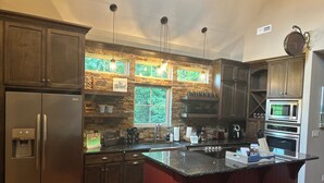 Eine moderne Küche mit einem Edelstahlkühlschrank, einem Einbauofen und einer Mikrowelle. Eine große Kochinsel mit Spüle und Fenster darüber. Die Küche verfügt über Holzschränke und eine Stein-Spritzschutzwand.