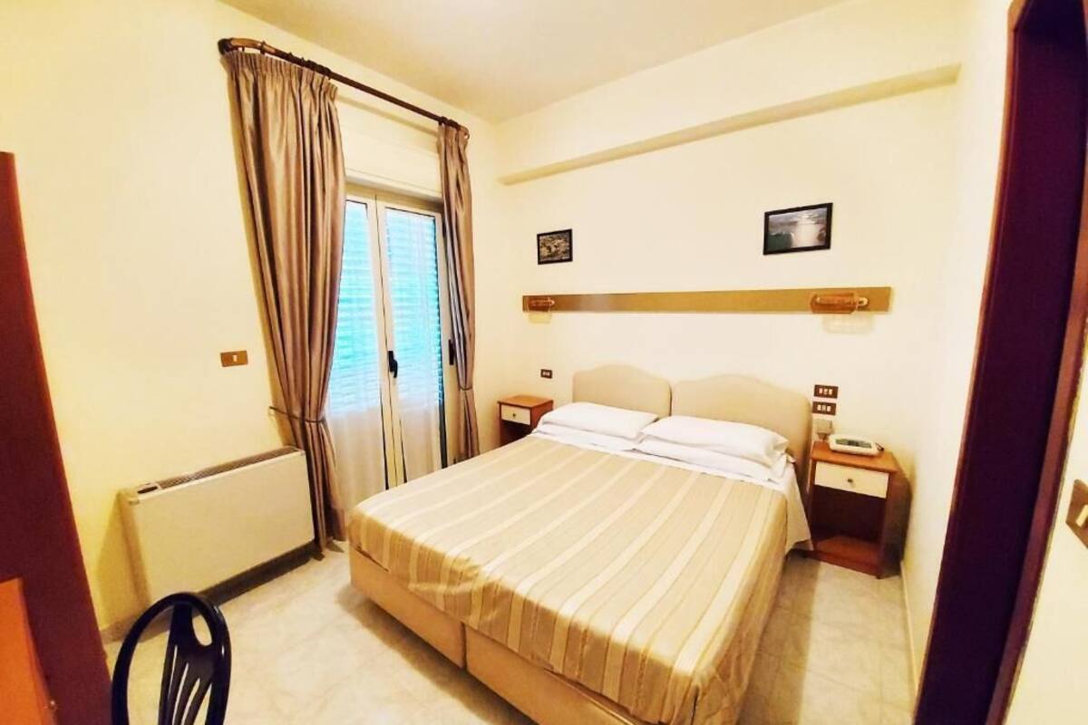 A hotel room with a bed, a chair, a nightstand, a bedside table, a radiator, and a window with curtains.
