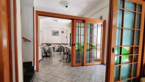 Um hall de hotel com piso de azulejos, área de refeições com mesas e cadeiras, e uma grande porta de madeira com painéis de vidro.