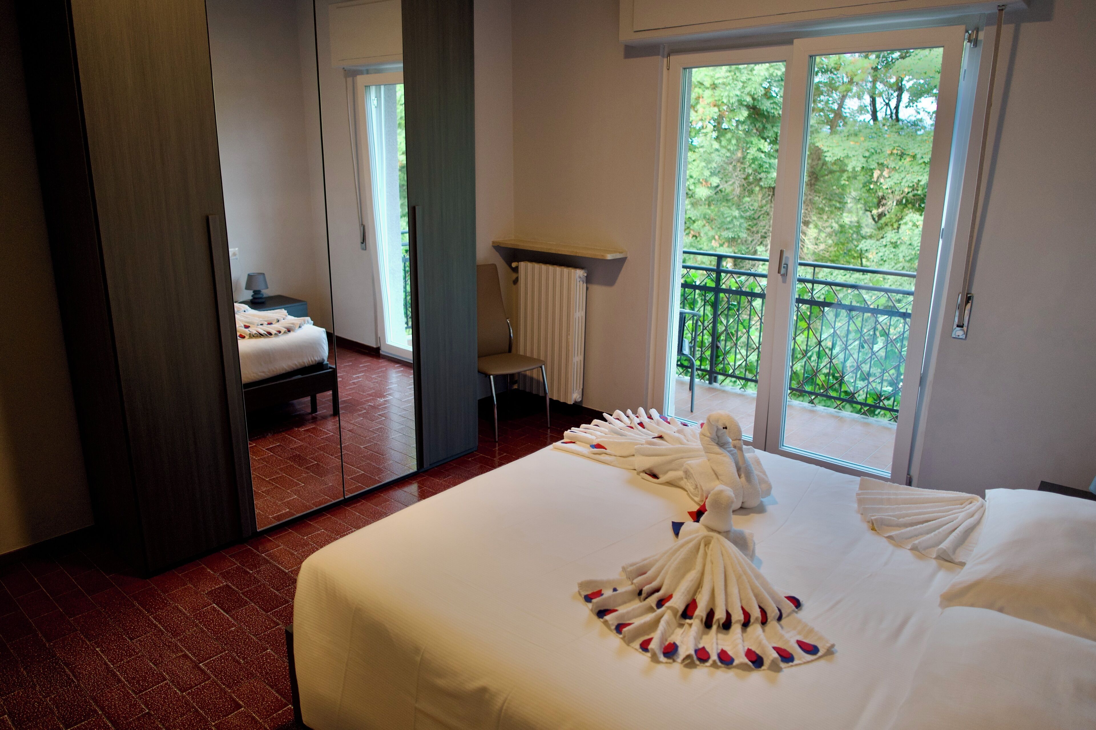 A hotel room with a large bed, a mirror, a chair, a small table, and a balcony with a view of trees.