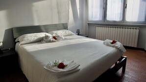 Ein ordentlich bezogenes Bett mit weißen Leinentüchern und Handtüchern in Rosenform, eine Nachttischlampe und ein Fenster mit Jalousien.