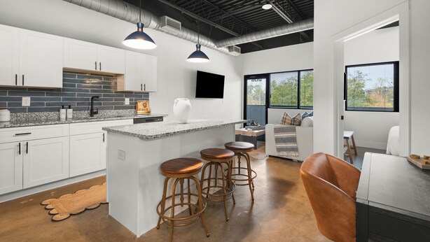 A modern kitchen with white cabinets, a granite countertop, and a central island with stools. There is a large window providing natural light and a view of greenery outside.