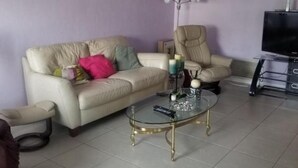 Una sala de estar con un sofá color beige, una mesa de centro de vidrio, un sillón color beige y un televisor sobre un mueble.