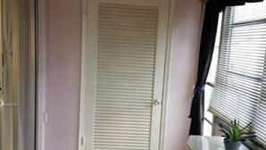 Una habitación con puerta corrediza de vidrio, una puerta blanca con persianas horizontales, piso de baldosas, un armario color crema con detalles decorativos y un ventanal con persianas.