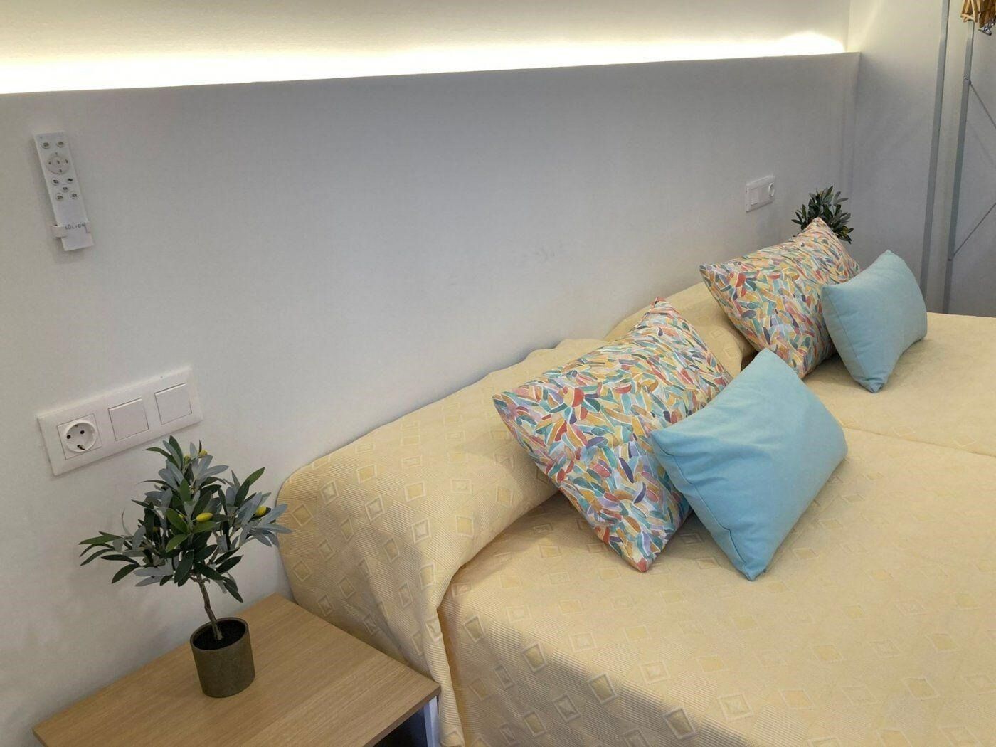 Una cama bien tendida con almohadones estampados, una planta en la mesita de noche y un enchufe de pared.
