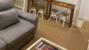 Ein Wohnzimmer mit grauem Sofa, einem Couchtisch mit Zeitschrift, einer Holzbank mit Hockern und einer Wendeltreppe an einer Steinwand.