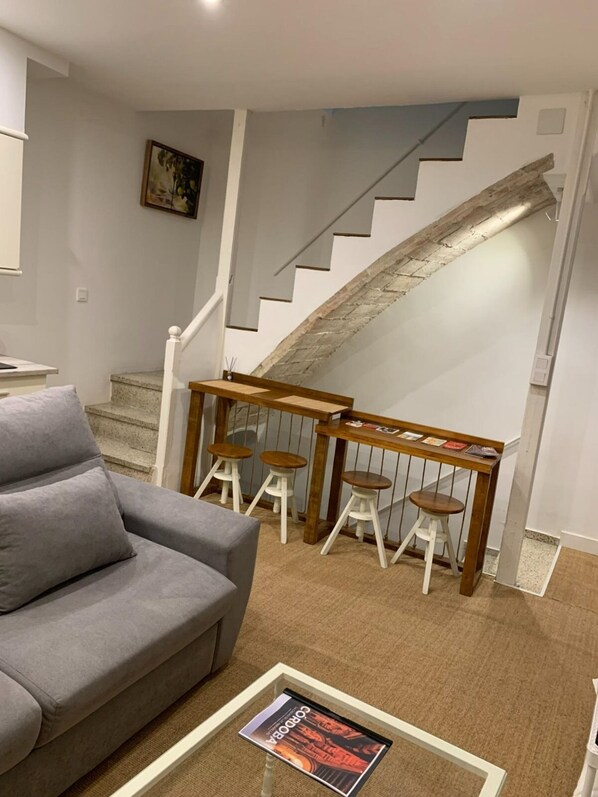 Una sala de estar con un sofá gris, una mesa de centro con una revista, un banco de madera con taburetes y una escalera con pared de piedra.