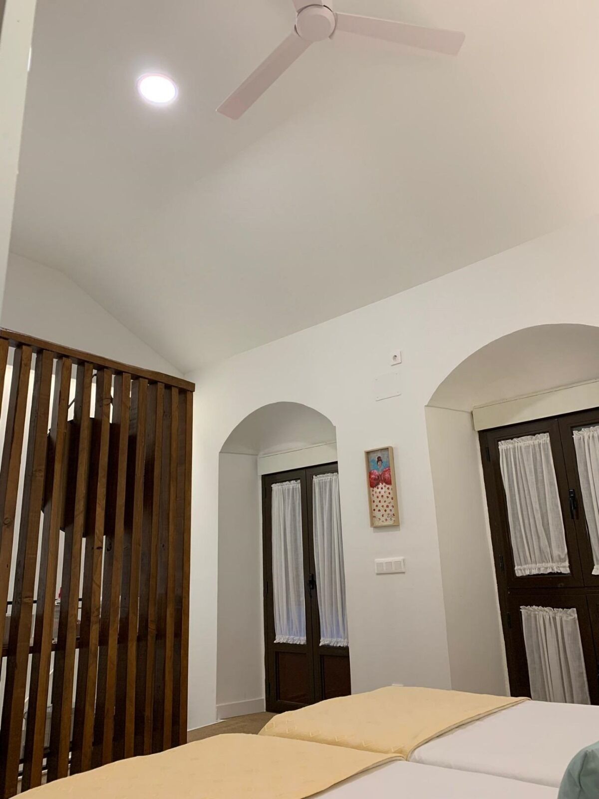 Ein Hotelzimmer mit Bett, Deckenventilator, Fenster mit Vorhängen und Raumteiler aus Holz.