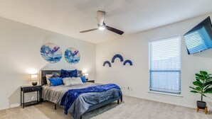 Een slaapkamer met een bed, nachtkastjes, een plafondventilator, wanddecoratie, een raam met jaloezieën en een potplant.