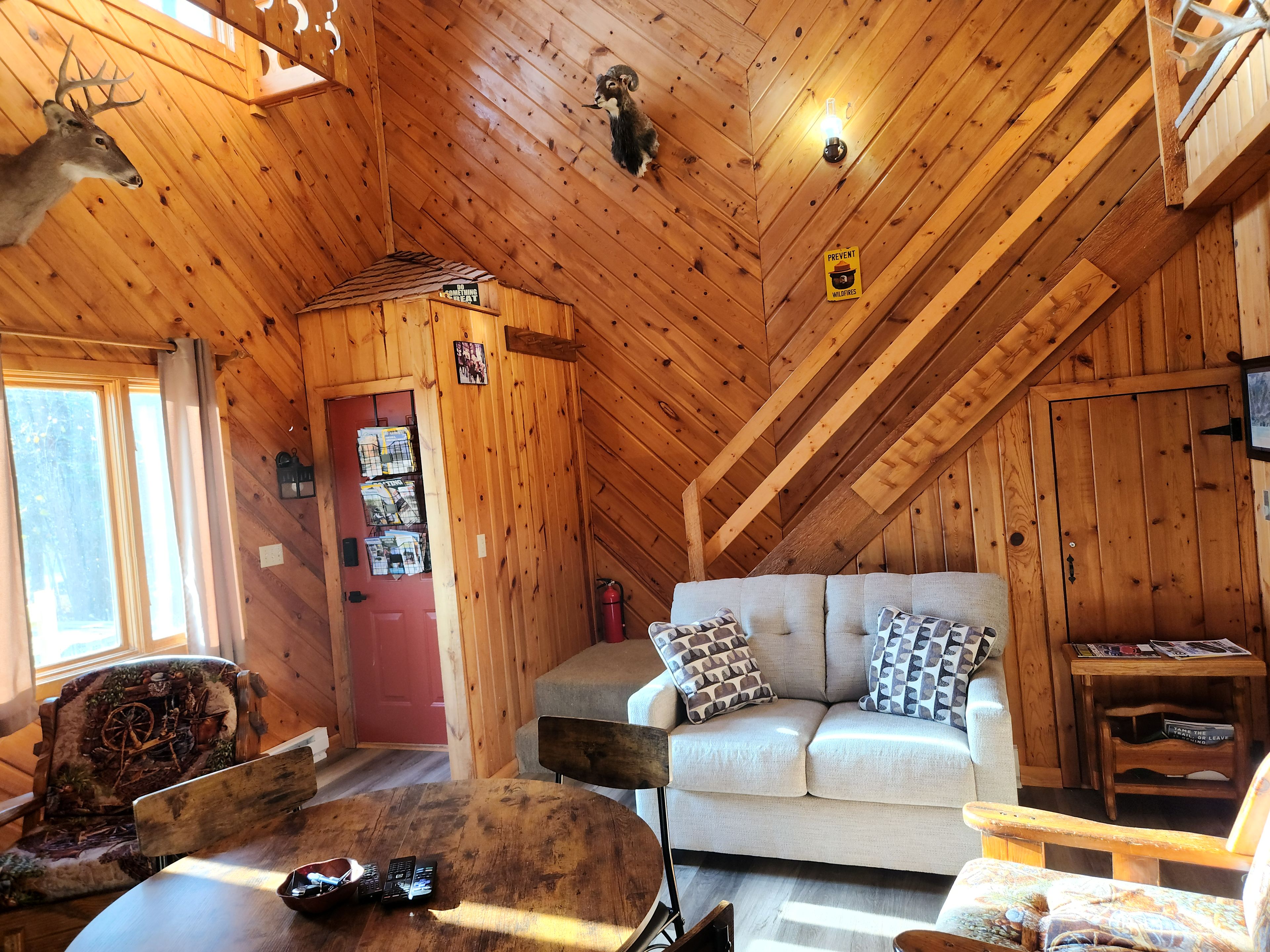 A cozy cabin interior with a wooden ceiling, a red door, a white sofa, and a rustic chair.